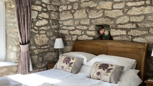 Un dormitorio con pared de piedra, cabecera de madera, cama con almohadas estampadas, lámpara de noche y ventana con cortinas.