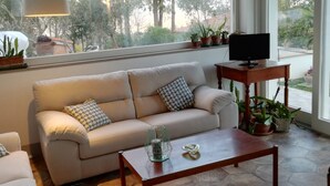 Uma sala de estar moderna com um sofá, uma mesa de centro e uma TV sobre um suporte de madeira. Há plantas em vasos no parapeito da janela e um abajur sobre a mesa.