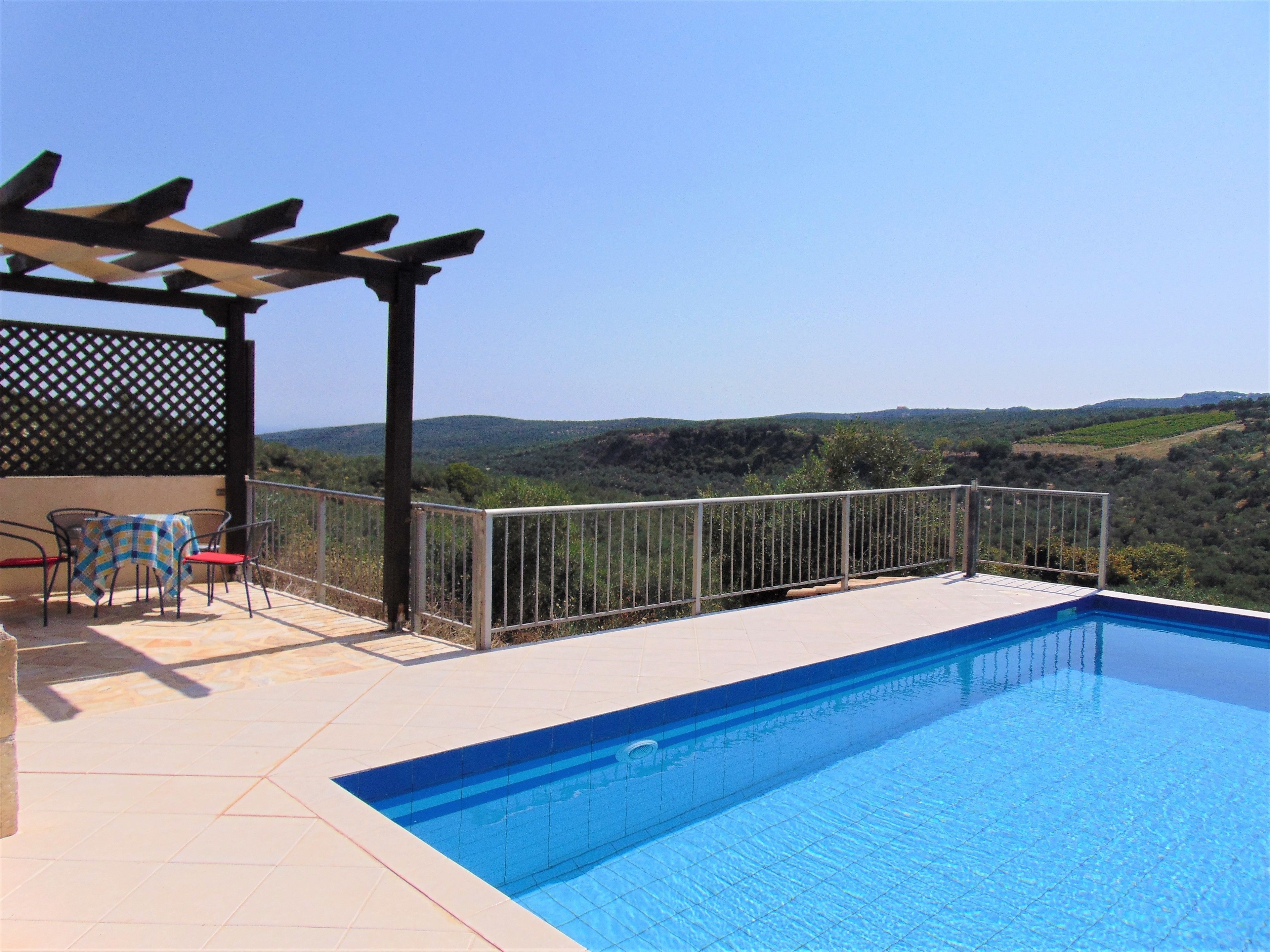 Piscina com pergolado e cadeiras, cercada por grade de metal, com vista para uma paisagem montanhosa.