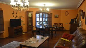 Ein Wohnzimmer mit Fernseher, einer Deckenleuchte, einem Esstisch mit Stühlen, einer Couch und einer Wanduhr.