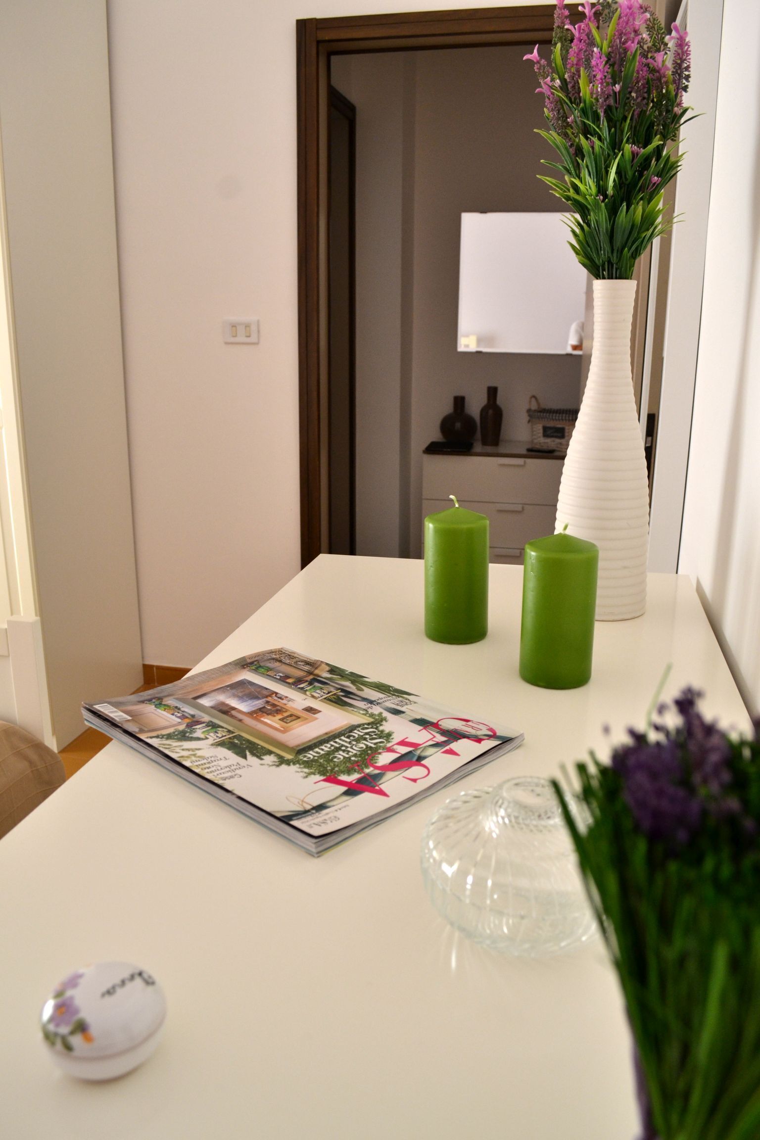 A white table with a magazine, two green candles, a clear glass bowl, and a white vase with purple flowers.