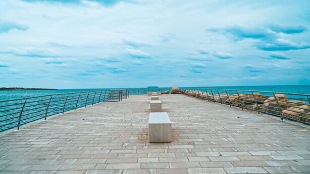 Une promenade dallée, bordée de garde-corps, qui mène à un plan d’eau.