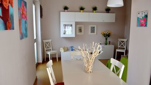 Un coin repas avec une table blanche, des chaises et un vase décoré de branches. Des plantes en pot sont disposées sur une étagère au-dessus de la table.