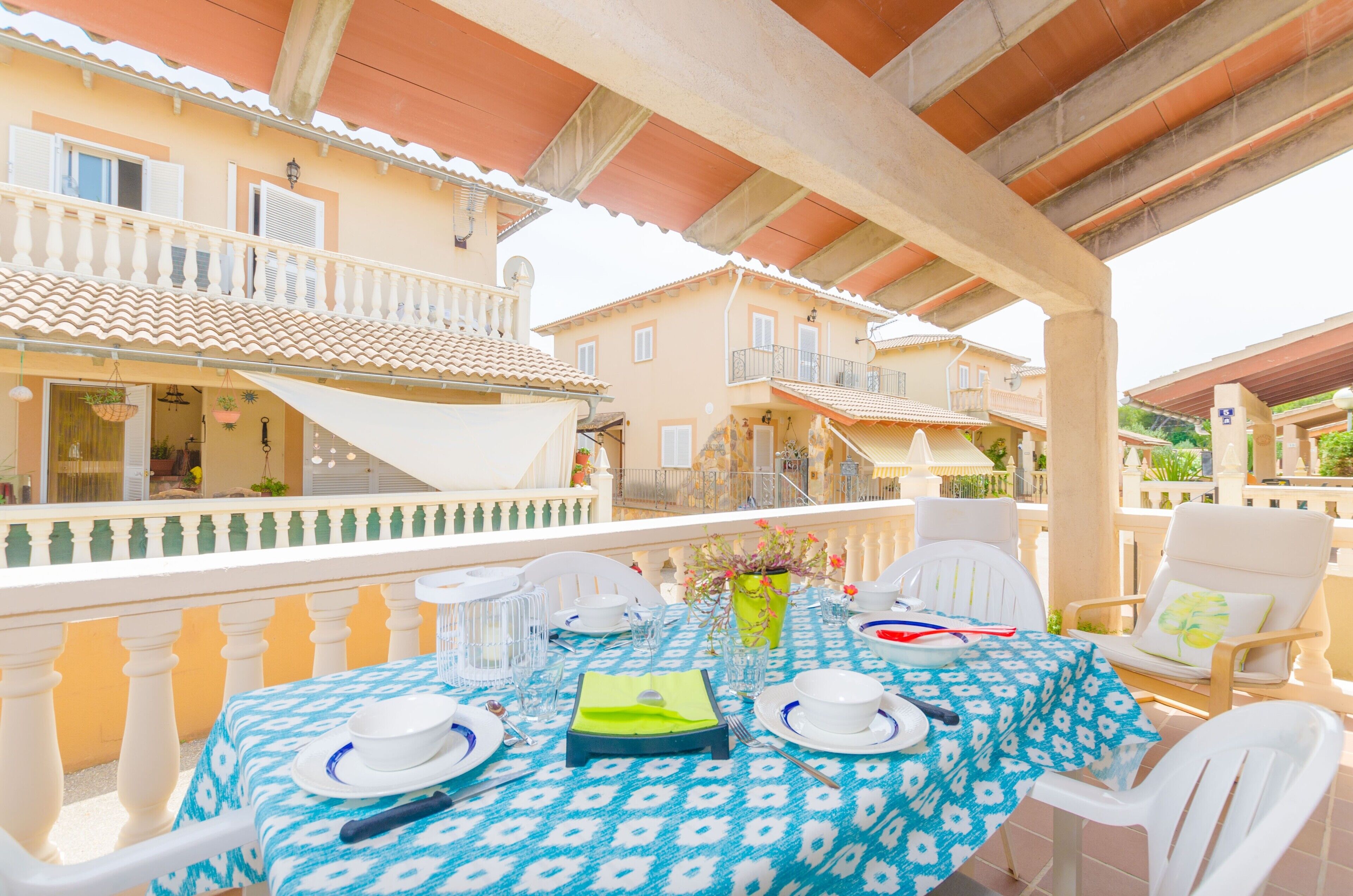 An outdoor dining area with a table set for two, white chairs, and a balcony overlooking a building with a tiled roof.