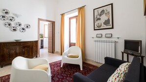 Un salón con un sofá, un sillón blanco, un tapete rojo estampado, un aparador de madera, un televisor y un cuadro enmarcado en la pared.