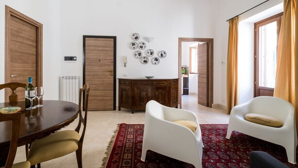 Un comedor con una mesa de madera, sillas y un tapete rojo estampado. Hay una silla blanca con cojín, otra silla blanca con cojín, y una silla negra. Se observa un mueble con encimera blanca al fondo.