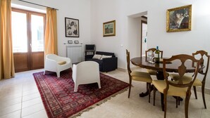 Una sala de estar con un sofá blanco, un sofá oscuro, una mesa de comedor con sillas, un tapete rojo estampado y un cuadro en la pared.