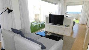 A modern living room with a grey sofa, a flat-screen TV on a white cabinet, and a view of a green lawn through large windows. - Sea Terrace (Socorro, El (Guimar))