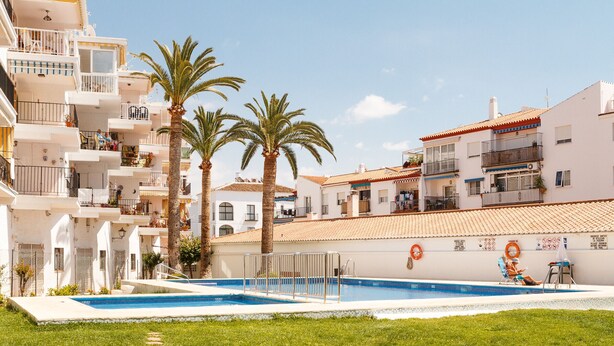 A swimming pool area with a clear sky, palm trees, and a building with balconies. - Coronado 129 Apartment by Casasol (Nerja)
