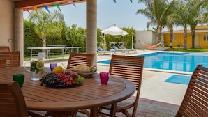 Een zwembadterras met een houten tafel, daarop fruit en een karaf, omringd door ligbedden en een hangmat.