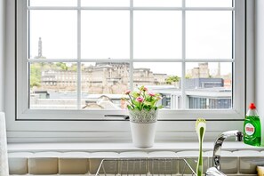 Una ventana con vistas a edificios, un jarrón blanco con flores, un cepillo de dientes y una botella de detergente.