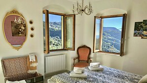 A bedroom with a large bed, a wooden chest, a chair, and a view of a mountainous landscape through two windows.