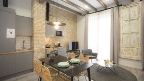 A modern kitchen with a brick wall, wooden beams, a dining area with wicker chairs, and a large window with curtains.