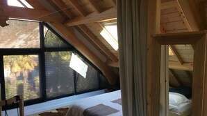 Un dormitorio con techo inclinado, una cama, una ventana con persianas y una estructura de techo de madera.