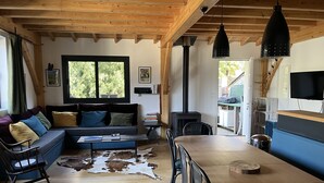 Una sala de estar acogedora con techo de madera, un sofá con cojines coloridos, una mesa de comedor con sillas, una chimenea y un televisor montado en la pared.