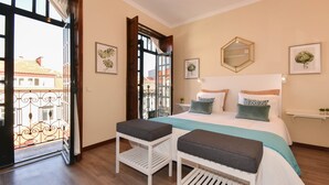 Un dormitorio con cama, dos mesitas de noche, una ventana con vistas a edificios y un balcón con barandilla de hierro forjado.
