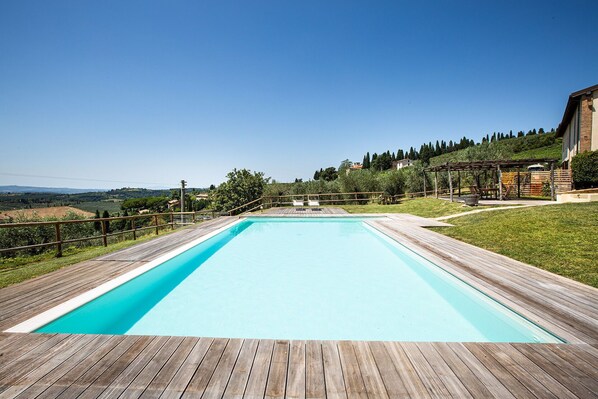 A rectangular swimming pool with a wooden deck surrounding it, set against a backdrop of a clear blue sky and a hilly landscape. - Beautiful apartment for 4 guests with A/C, pool, WIFI and TV, close to San Gimignano (Poggibonsi)
