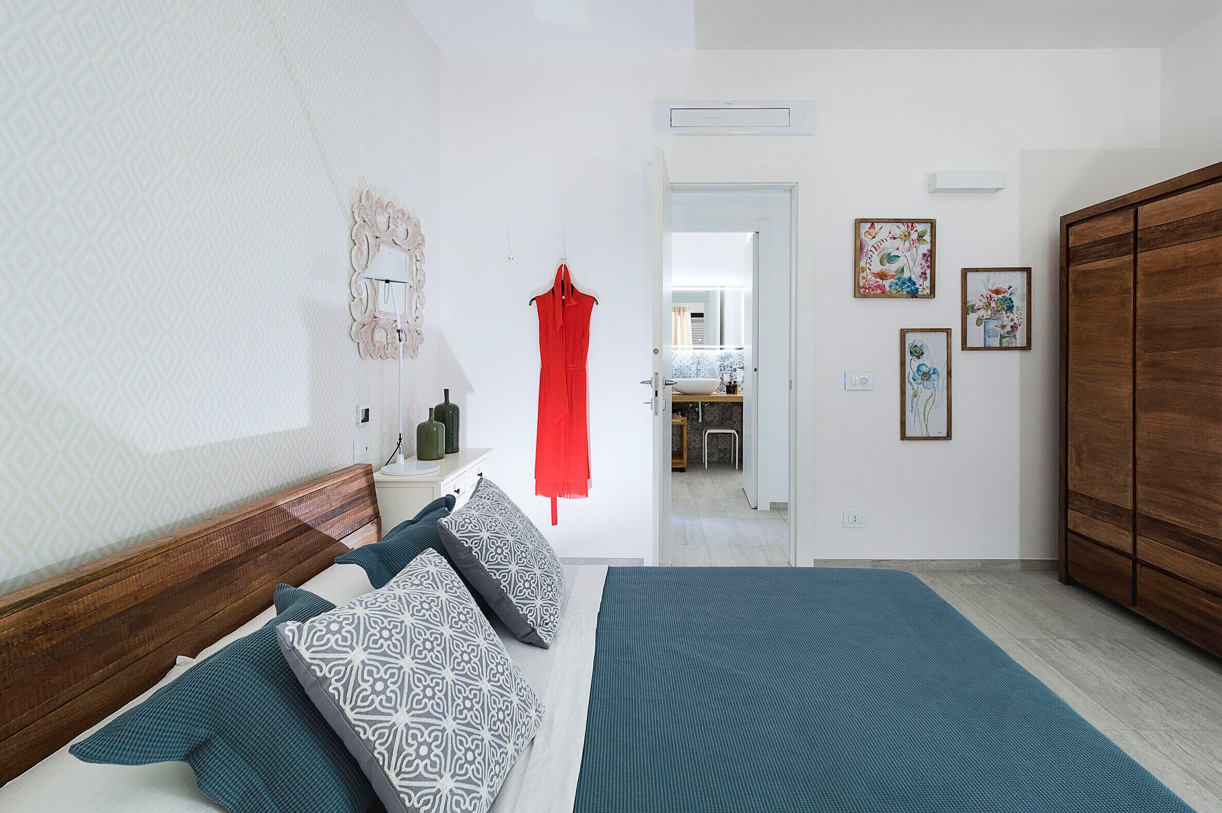 A bedroom with a wooden headboard, patterned pillows, a blue bedspread, and a red garment hanging on the wall.
