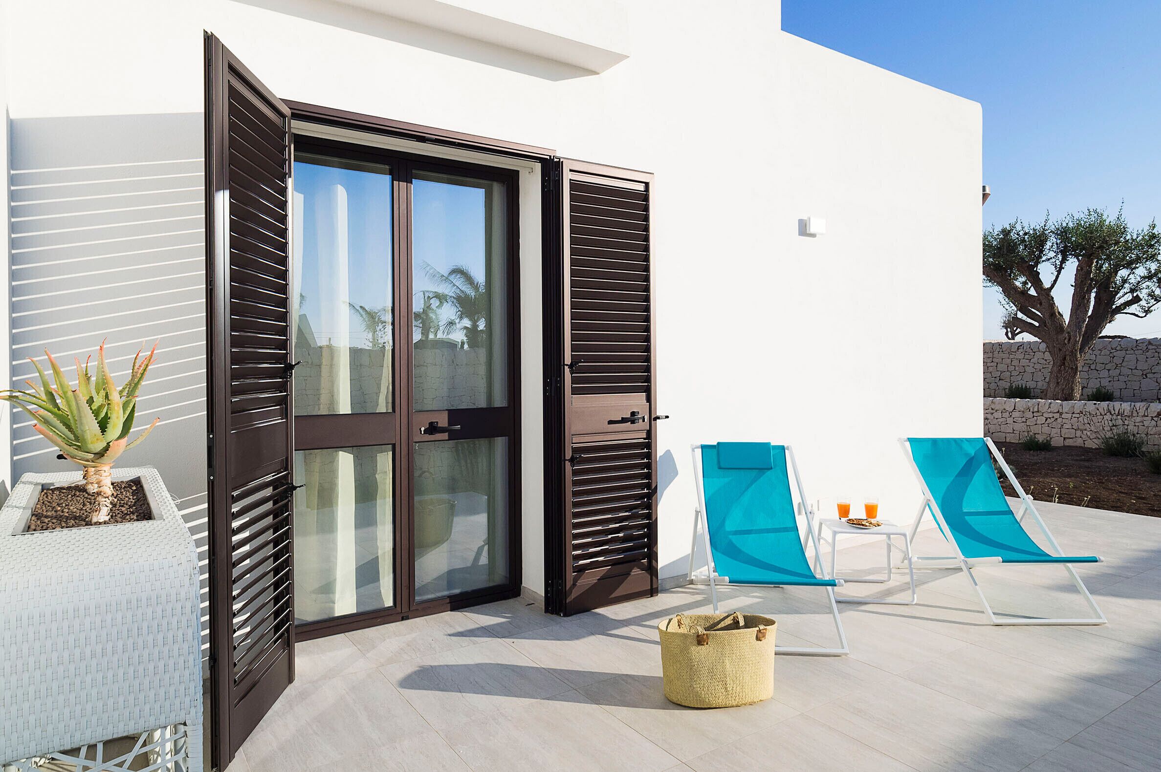 A modern outdoor area with a sliding glass door, two blue lounge chairs, a small table with drinks, and a potted plant.