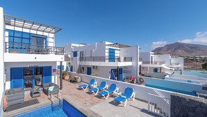 Un espace aménagé au bord de la piscine, avec des chaises longues, une piscine et un bâtiment aux volets bleus.