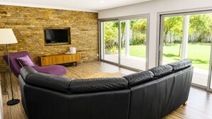 Een moderne woonkamer met een hoekbank, een flatscreen televisie aan een stenen muur, een houten kast en een grote glazen schuindeur die uitkomt op een tuin.