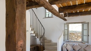 Una escalera con techo de vigas de madera, una pared de piedra visible a través de una ventana y un sofá beige con cojines.