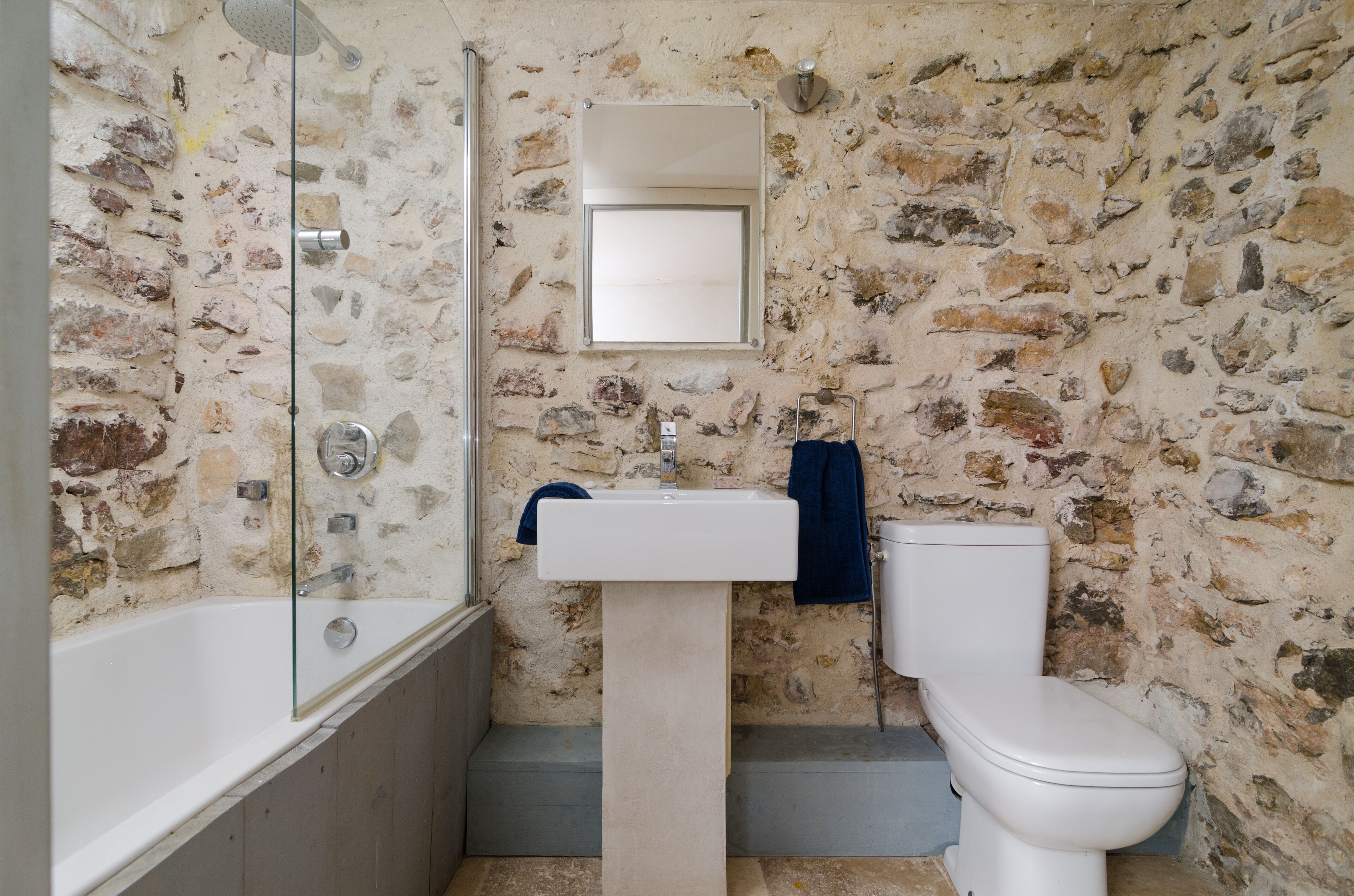 Un baño con pared de piedra, un lavamanos blanco, un inodoro blanco y una ducha con mampara de vidrio.