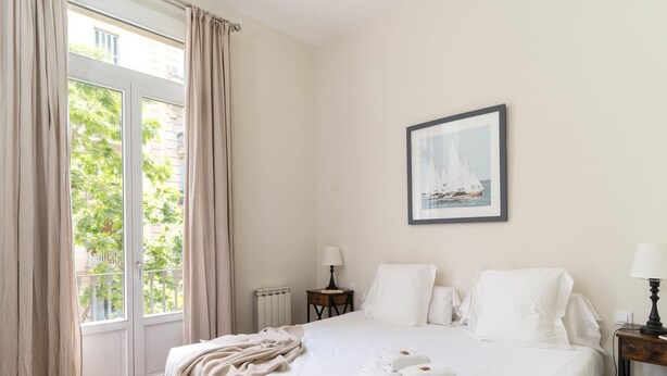 Ein Hotelzimmer mit einem großen Bett, einer Sitzbank, einem Fenster mit Vorhängen, einem gerahmten Bild an der Wand und einem Nachttisch mit einer Lampe.