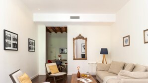 Ein Wohnzimmer mit Couch in Beige, einem Holztisch und einem Sessel mit gelber Kissen. An der Wand hängt ein Spiegel und ein Lampenstativ steht bereit.