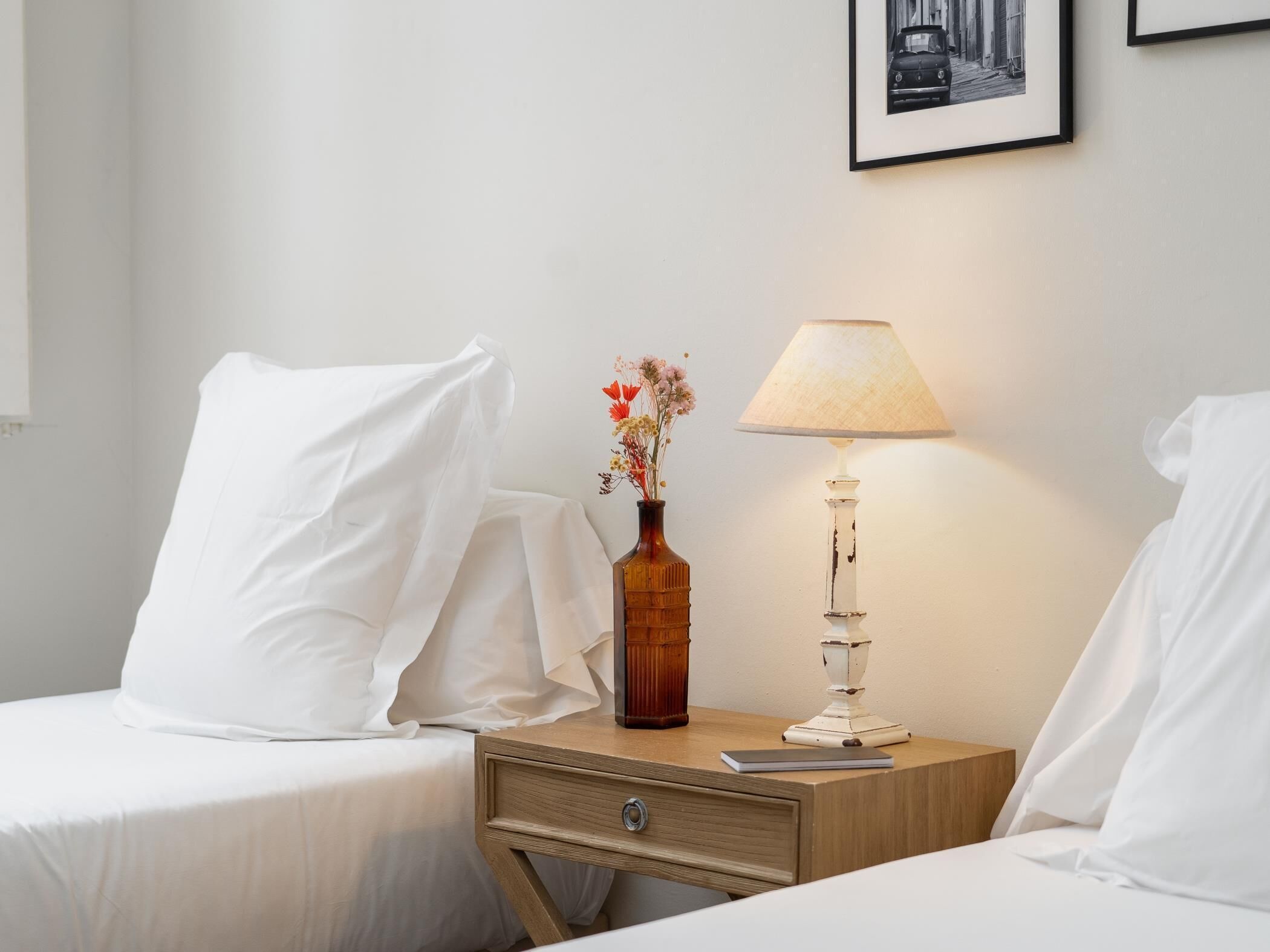 Un lit bien fait, avec du linge de lit blanc, une petite table de chevet en bois sur laquelle se trouvent une lampe et un vase rempli de fleurs, et un tableau encadré accroché au mur.