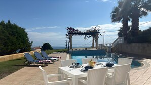 Een zwembadterras met een eettafel, lounge stoelen en een pergola met uitzicht op zee.
