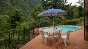Een zwembadterras met een tafel en stoelen, een blauwe parasol en uitzicht op een beboste heuvel.