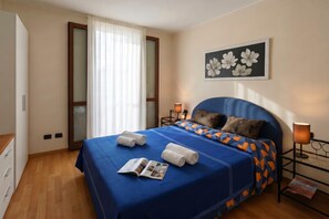 Ein Schlafzimmer mit einem Bett, einem Nachttisch, einer Lampe, einem Fenster mit Vorhängen und einer Wand mit einem Blumenbild.