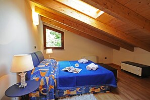 A bedroom with a sloped ceiling, a bed with a blue bedspread, a bedside table with a lamp, a dresser, and a window with a view of greenery.