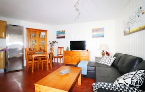 Een woonkamer met een bank, een televisie, een eettafel en een keuken met een koelkast.