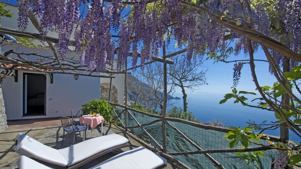 A balcony with white lounge chairs, a table with a pink tablecloth, and purple wisteria hanging above. - White Daisy is a small villa on two floors (Praiano)