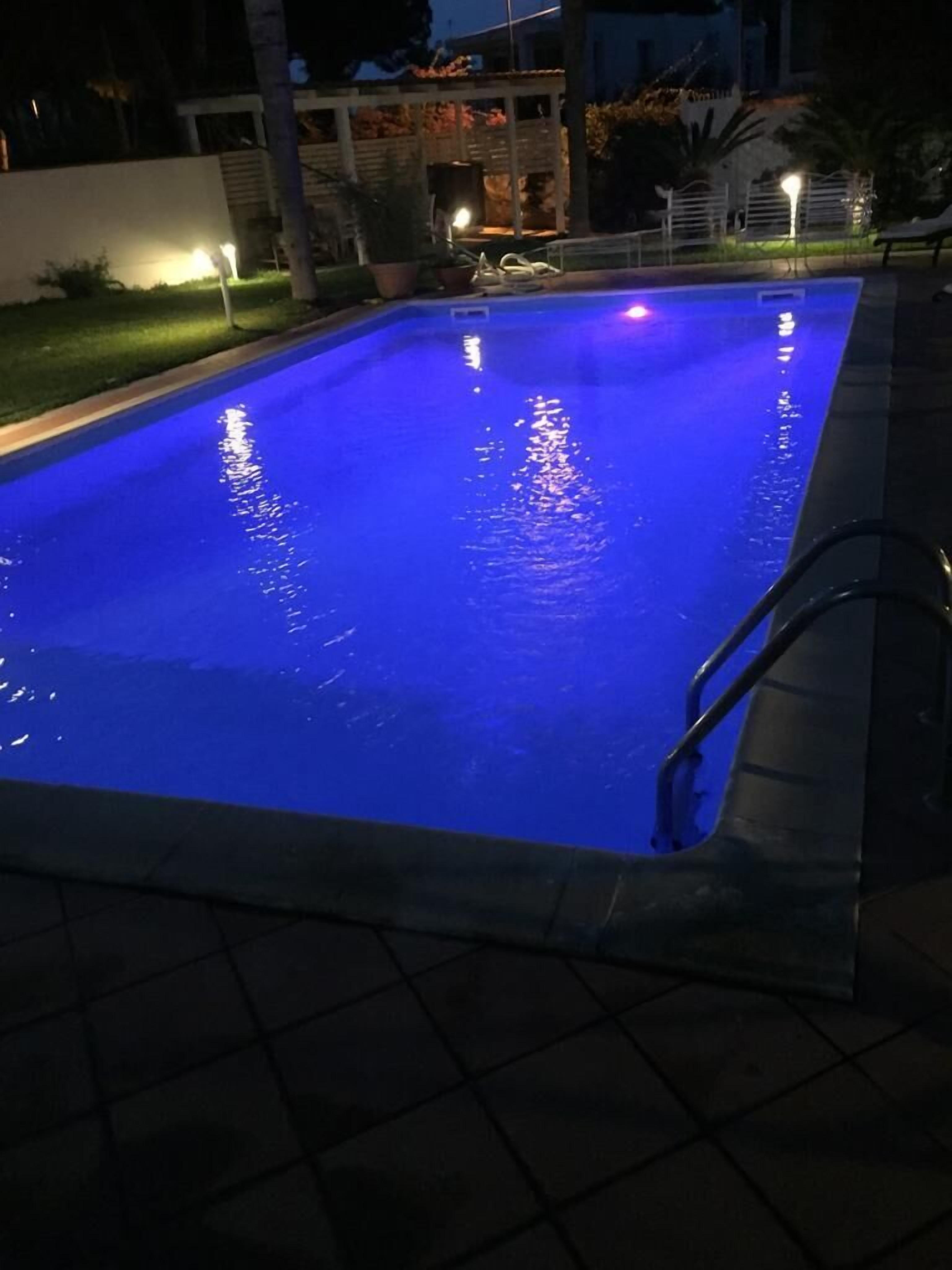 Piscina rectangular con iluminación azul, rodeada por un solárium empedrado y un muro bajo.