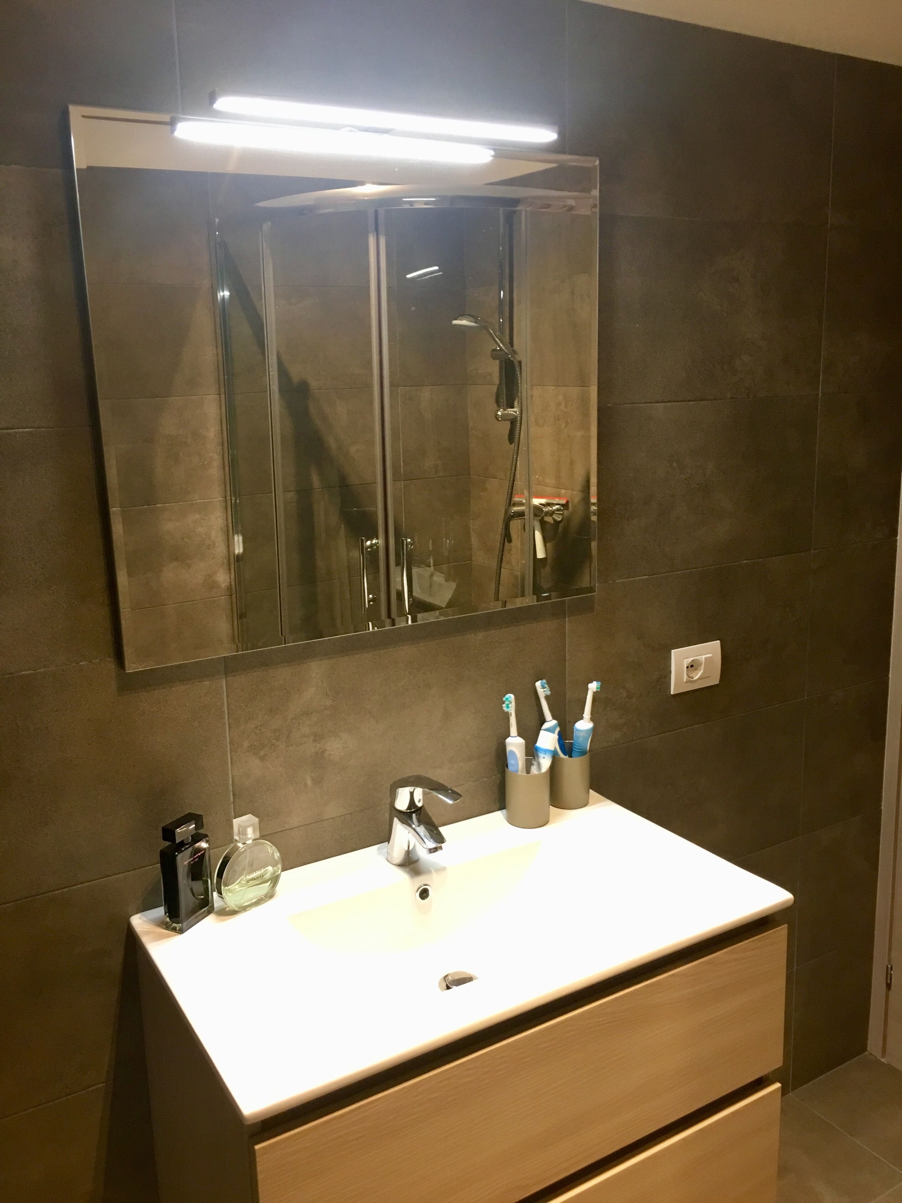 A bathroom with a white sink, a mirror, and a shower. There are toothbrushes in a holder and a bottle of hand soap on the sink.