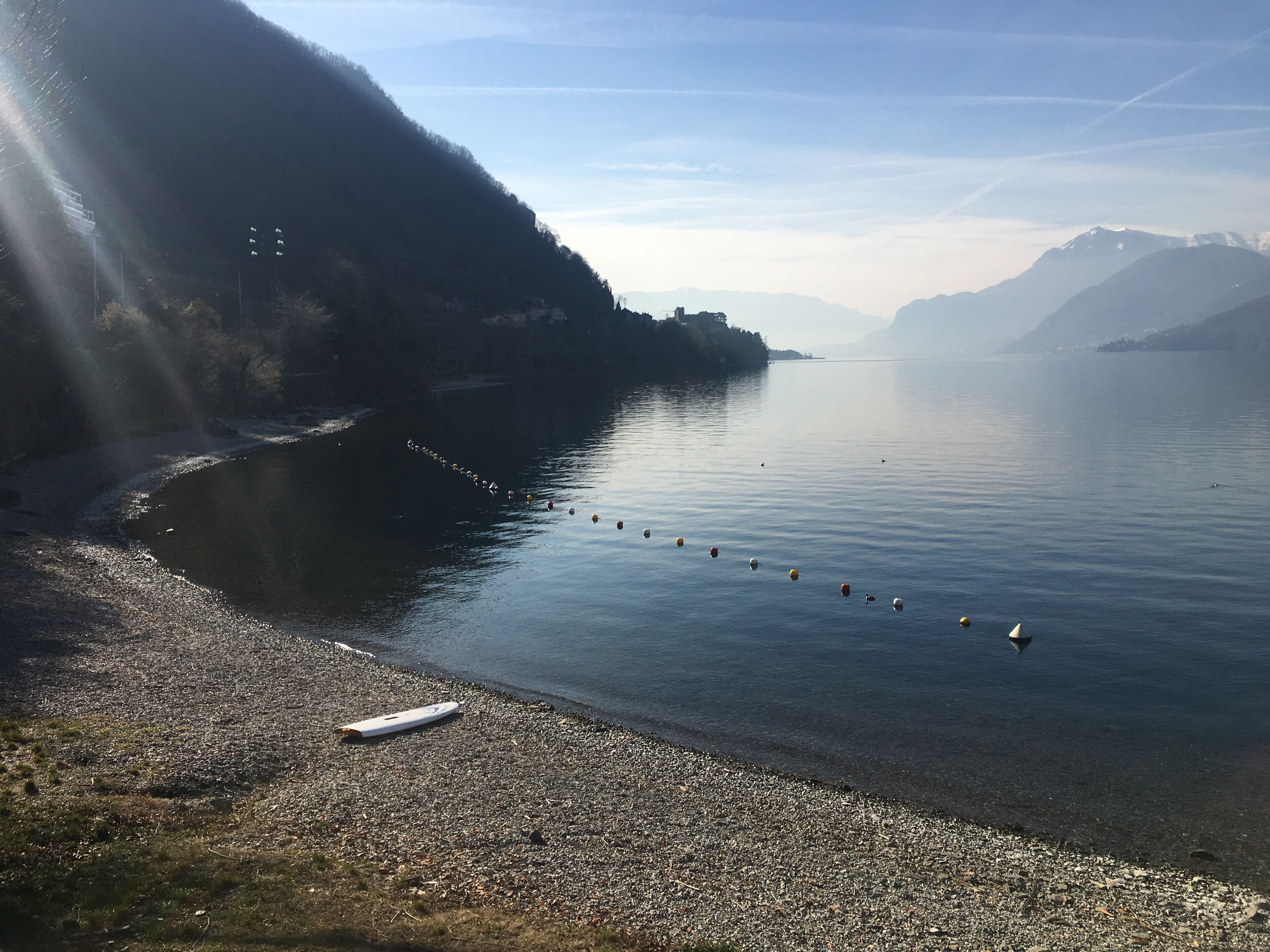 A calm lake with a row of buoys, a pebble beach, and a mountain in the background.