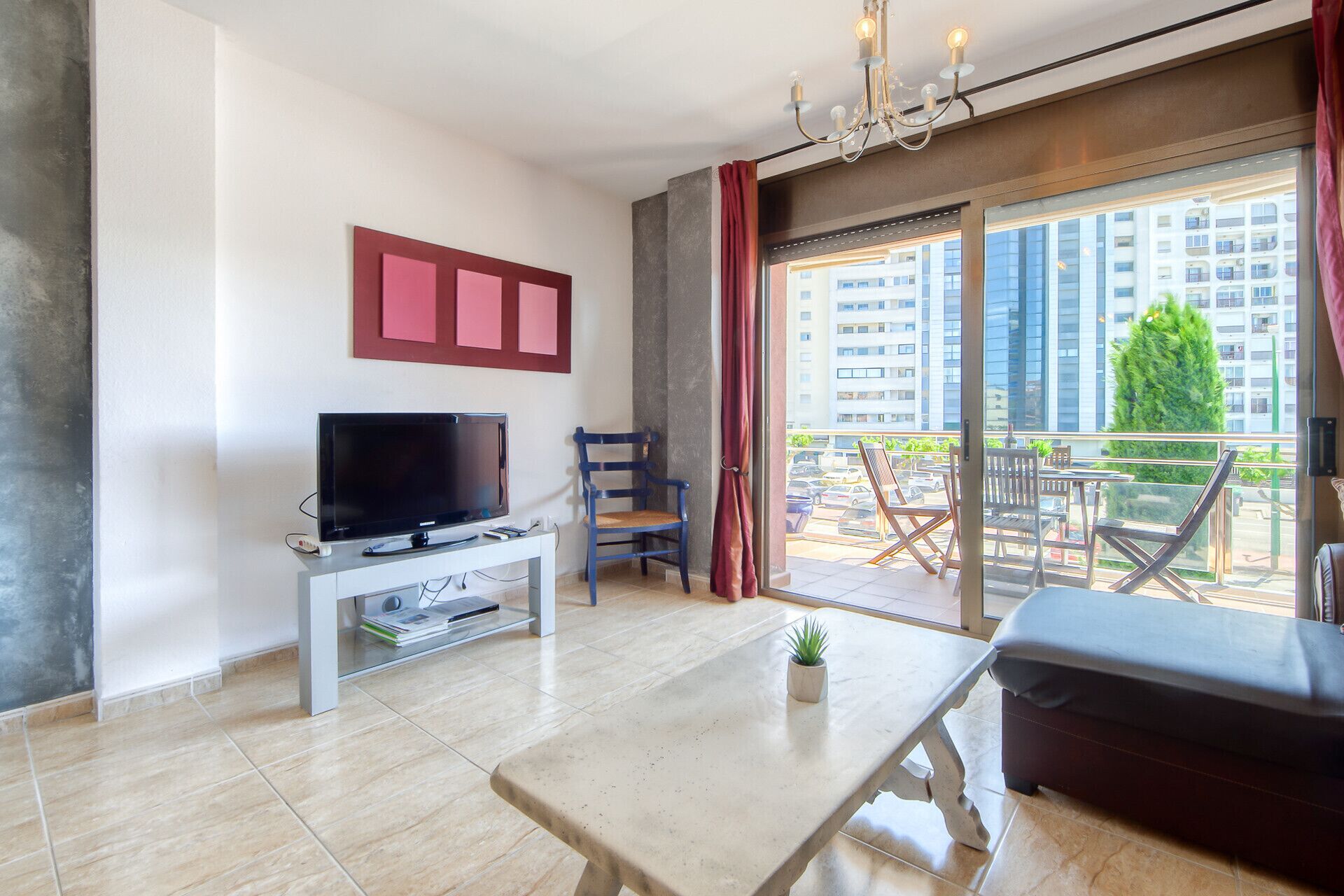 Un salon moderne avec un téléviseur à écran plat posé sur une console blanche, une table à manger et un canapé. Une grande fenêtre offre une vue sur le paysage urbain et la verdure.