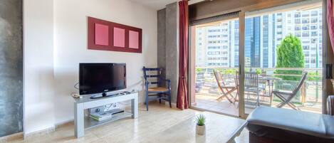 Un salon moderne avec un téléviseur à écran plat posé sur une console blanche, une table à manger et un canapé. Une grande fenêtre offre une vue sur le paysage urbain et la verdure.