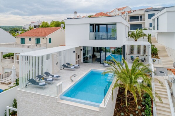 A modern villa with a swimming pool, outdoor seating, and a view of neighboring buildings.