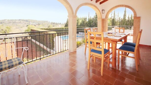A balcony with a wooden table and chairs, a view of the countryside, and a pool in the background.