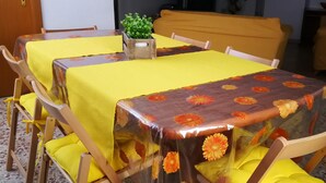 A dining area with a table covered in yellow and brown floral tablecloths, wooden chairs with yellow cushions, and a small potted plant on the table.
