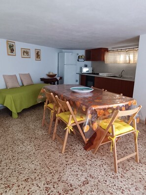 A compact living space with a dining area featuring a table covered with a floral tablecloth, wooden chairs with yellow cushions, a green bed, and a kitchenette in the background. - Julia House. Only 250 m from the beach and promenade location of Montalbano (Donnalucata)