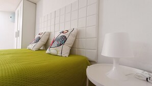 A hotel room with a green bedspread, two decorative pillows with a floral pattern, a white headboard, and a bedside table with a white lamp and a power outlet.