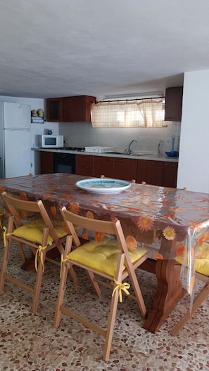 A dining area with a table covered by a floral tablecloth, wooden chairs with yellow cushions, a white refrigerator, a microwave, and a sink with a faucet. - Julia House. Only 250 m from the beach and promenade location of Montalbano (Donnalucata)