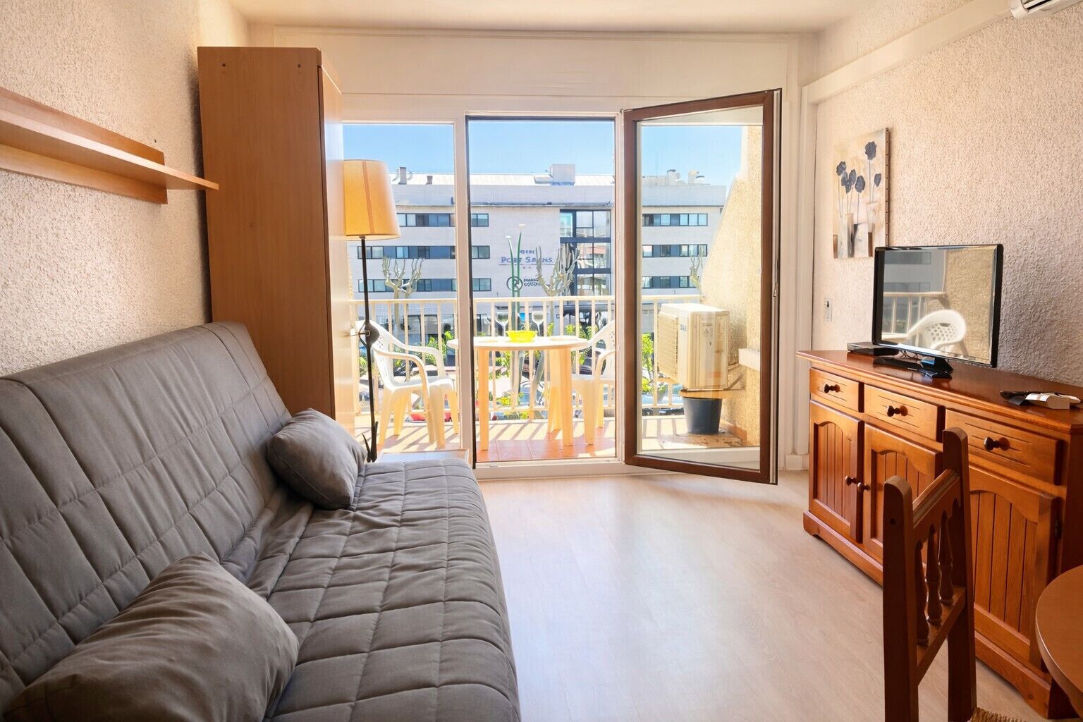 Ein Zimmer mit grauem Sofa, einem Holzkasten und einem Balkon mit Tisch und Stühlen.