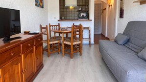 A living room with a sofa, a TV, a dining table with chairs, and a kitchen area with a window and a hanging light fixture. - 0167-POBLAT TIPIC Studio near the beach (Empuriabrava)