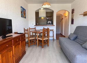 Un soggiorno con divano, televisore, tavolo da pranzo con sedie e angolo cottura con finestra e lampadario.